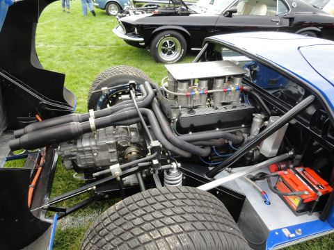 1964 Ford Fairlane - The engine bay of a blue race car is shown, with a black muscle car visible behind it on a grassy field.