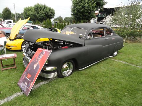 1964 Ford Fairlane - A black sedan is displayed with its hood open at a car show, a movie poster. Other vehicles are present in the background.