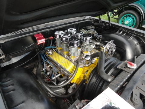 1964 Ford Fairlane - Engine bay view of a V8 with multiple carburetors, with another vehicle partially visible in the background.