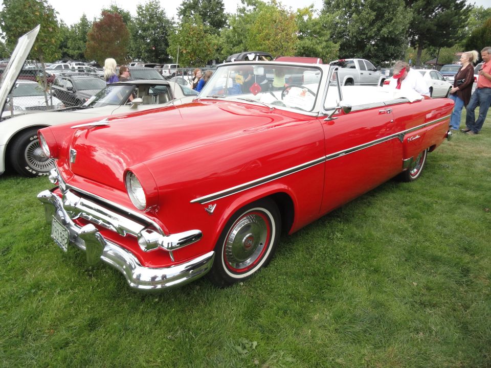 Red 1955 Ford Fairlane convertible parked on grass with other cars in background