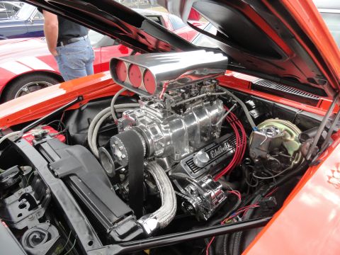 1964 Ford Fairlane - Engine bay of an orange car with a prominent supercharger and other chrome components is visible, with other vehicles in the background.