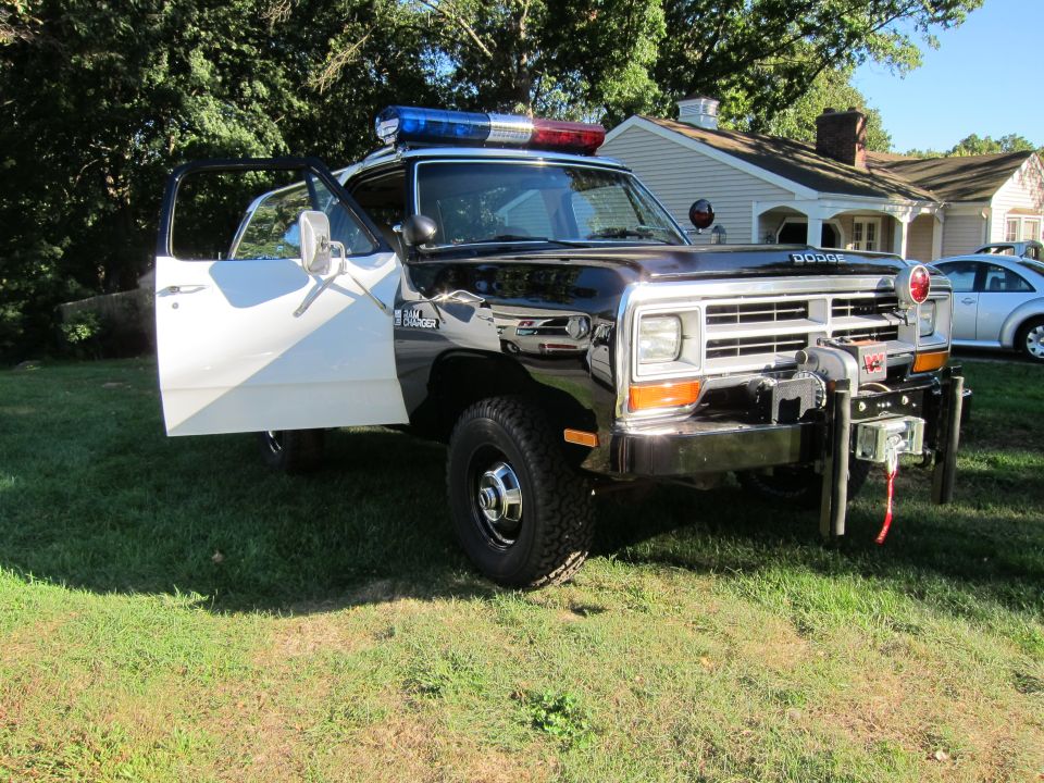 1987 Dodge 1500 police truck with open driver's door, light bar on roof, and winch on front bumper. Silver car parked in background