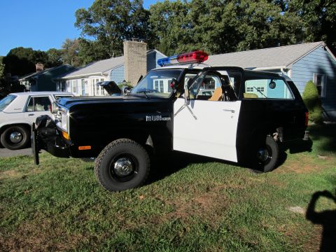 1987 Dodge 1500 - A two-tone police SUV is parked on a grassy lawn in front of houses.