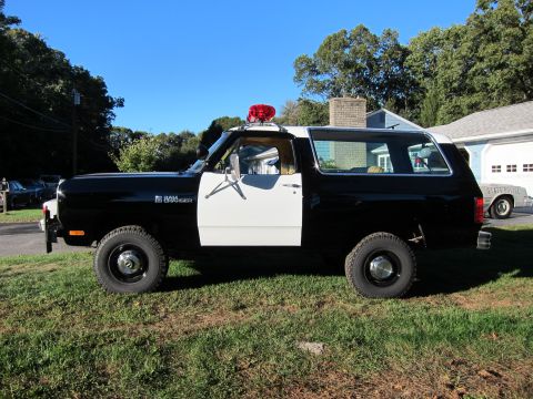 1987 Dodge 1500 - A two-tone police SUV, likely a Ramcharger, is parked on a grassy area with a house in the background. Another vehicle marked 'STATE POLICE…