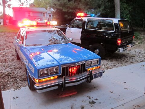 1987 Dodge 1500 - Two police vehicles, one a sedan and the other an SUV, are parked side-by-side with their emergency lights illuminated.