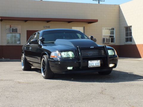 2003 Ford Crown Victoria - Crown Vic with push bar and EVIL VIC plate in a lot beside a tan industrial building.