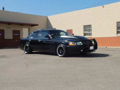 2003 Ford Crown Victoria - Crown Vic front-corner profile on black wheels beside a tan warehouse building.