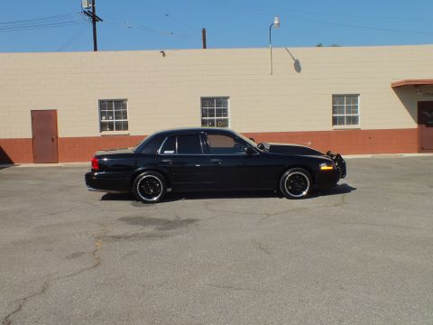2003 Ford Crown Victoria - Full side profile of a blacked-out Crown Vic on black wheels by a beige wall.