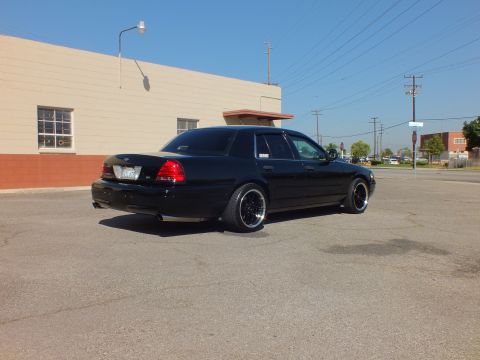 2003 Ford Crown Victoria - Rear quarter of a blacked-out Crown Victoria sitting low on dark machined-lip wheels against an industrial backdrop.