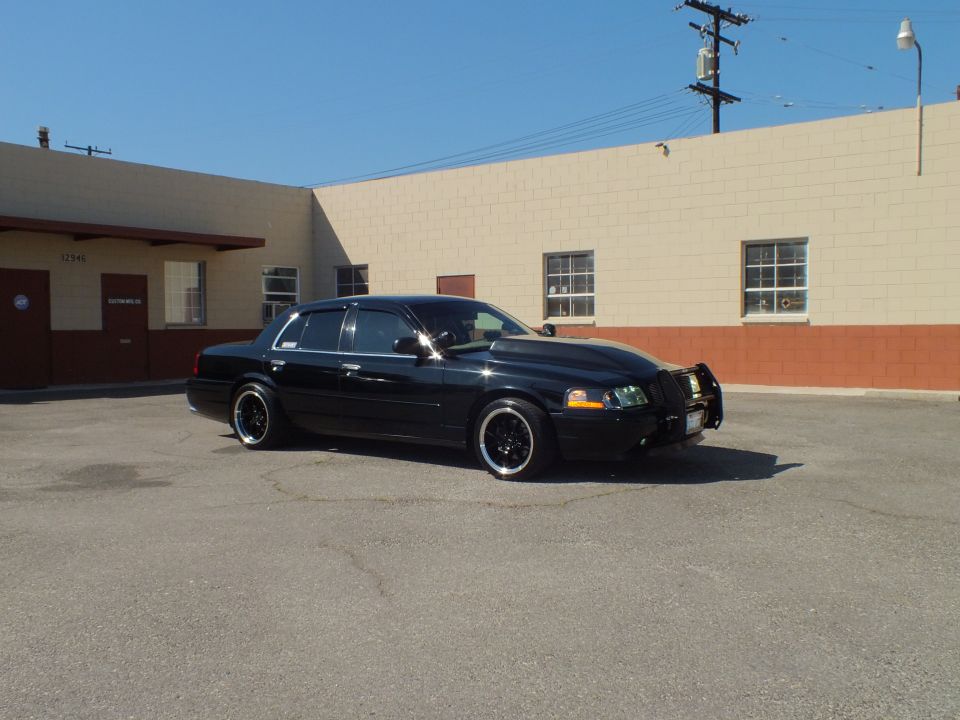 Black Crown Victoria front quarter, grille guard, black wheels, beige building behind