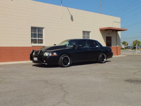2003 Ford Crown Victoria - Black Crown Victoria with grille guard parked low along a tan building with a palm tree behind.