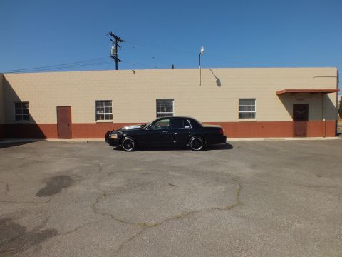 2003 Ford Crown Victoria - Distant side view of a black Ford sedan against a tan industrial wall under clear blue sky.