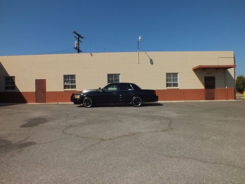 2003 Ford Crown Victoria - Wide shot of a black Ford sedan parked across an empty lot beneath a warehouse wall.