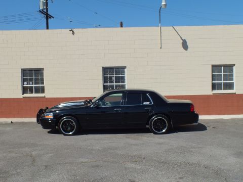 2003 Ford Crown Victoria - Side profile of a black Ford sedan with grille guard against a tan block building.