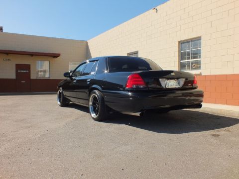 2003 Ford Crown Victoria - Low rear angle of a glossy black Ford sedan with dual tailpipes beside a block wall.