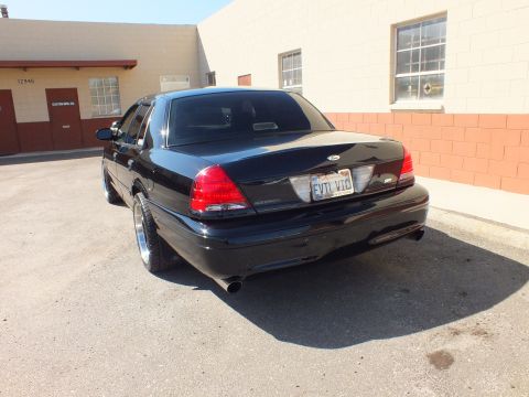 2003 Ford Crown Victoria - Rear three-quarter of a black Ford sedan with EVIL VIC plate beside a warehouse wall.