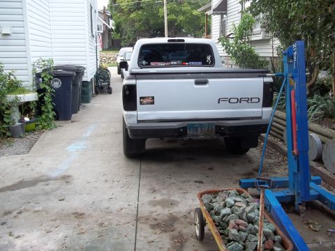 1970 Ford Ranchero - A white Ford pickup truck is parked in a driveway next to a house, with a blue engine hoist and a wheelbarrow visible.