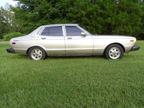 1978 Nissan Maxima - A silver 1978 Nissan Maxima sedan is shown from the side, parked on a grassy area with trees in the background.