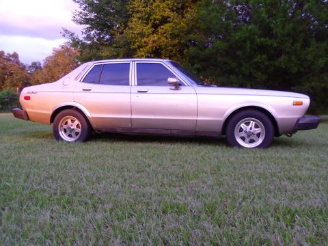1978 Nissan Maxima - This 1978 Nissan Maxima sedan is seen from the side, parked on a grassy area with trees in the background.