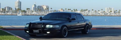 2003 Ford Crown Victoria - Black Crown Victoria with brush guard posed by a harbor, skyscrapers and sailboats in the distance