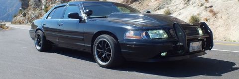 2003 Ford Crown Victoria - Front three-quarter of a black Crown Victoria with push bar and lit headlights on a hill road