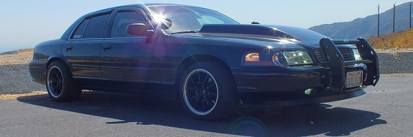 Black Ford Crown Victoria with push bar and black wheels on mountain road with sun glare