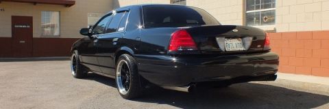 2003 Ford Crown Victoria - Rear quarter of a lowered black sedan parked by a brick building, aftermarket dark wheels and dual exhaust tip