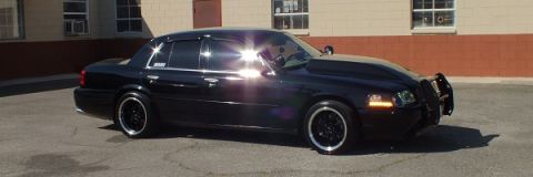 2003 Ford Crown Victoria - Profile of a black sedan parked by a tan building, black wheels and front push bar, sun reflecting off paint