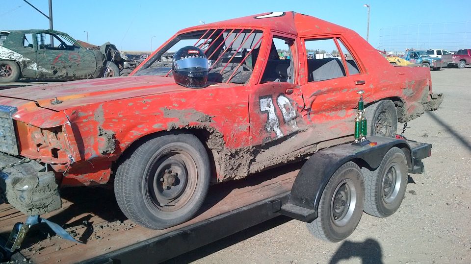 Mud-covered red derby sedan with roll cage and helmet loaded on a flatbed trailer