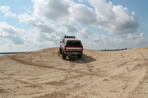1990 Ford Bronco - SUV with off-road modifications navigates a sandy terrain under a cloudy sky.