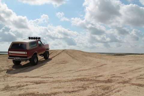 1990 Ford Bronco - This lifted SUV is shown from the rear as it climbs a sandy incline under a cloudy sky.