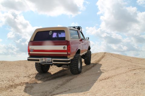1990 Ford Bronco - This lifted 1990 Ford Bronco navigates a sandy incline under a partly cloudy sky.