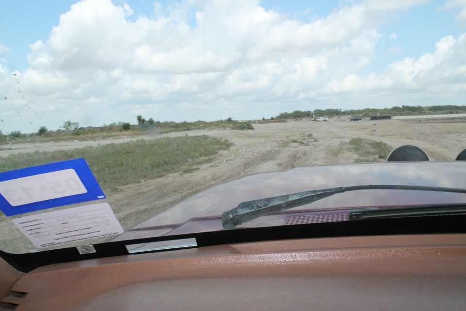 Interior view from a vehicle, showing dashboard and windshield looking out at a dirt road with other vehicles in the distance