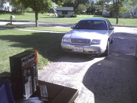 2003 Ford Crown Victoria - A silver sedan is parked in a driveway with a box of oil and cleaner on a table nearby.
