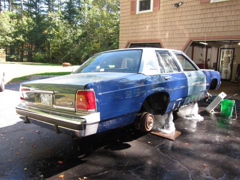 1991 Ford LTD Crown Victoria - A blue and silver sedan is elevated on blocks in a driveway, undergoing repairs or restoration.