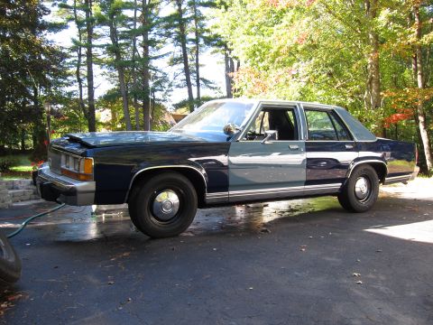 1991 Ford LTD Crown Victoria - A two-tone sedan with its hood partially open sits on a wet asphalt surface.