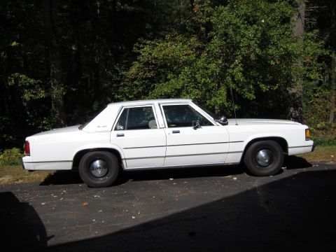 1991 Ford LTD Crown Victoria - A white sedan is parked on an asphalt surface with trees in the background.