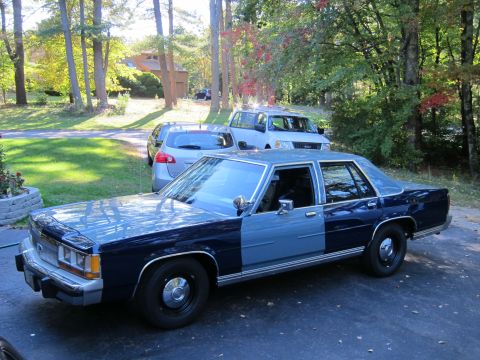 1991 Ford LTD Crown Victoria - A two-tone blue 1991 Ford Crown Victoria LTD sedan is parked in a driveway with other vehicles and trees in the background.