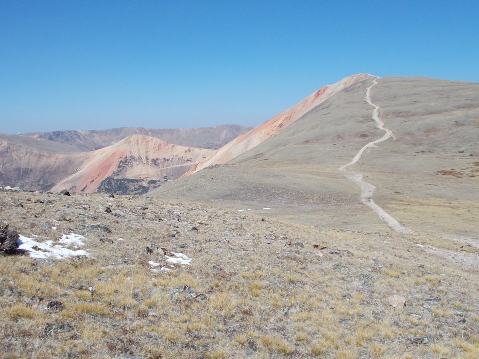 Mountain landscape with a winding dirt path up a hill under a clear blue sky. Some dry grass and patches of snow are visible in the foreground