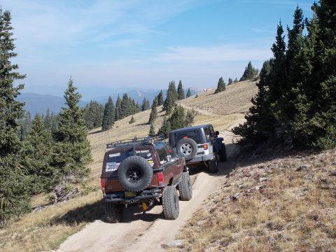 1986 Ford Bronco II - Two off-road vehicles navigate a steep, dusty trail through a mountainous, forested landscape under a clear blue sky.