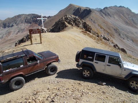 1986 Ford Bronco II - Two off-road vehicles are parked on a mountain trail near a summit marker and a large cross.