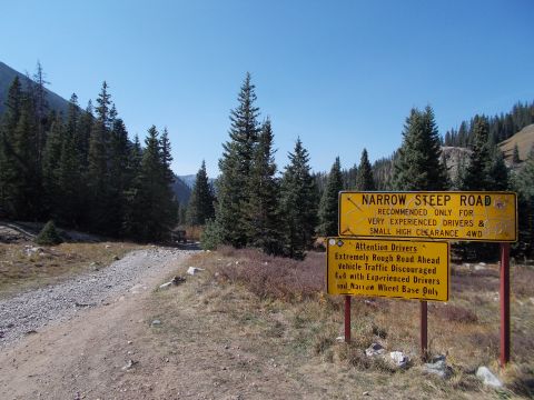 1986 Ford Bronco II - An off-road vehicle navigates a dirt road in a mountainous, forested area, passing by warning signs for a narrow and steep road.