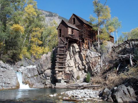 1986 Ford Bronco II - An old wooden mill building precariously perched on a cliff overlooking a waterfall and river, surrounded by autumn trees.