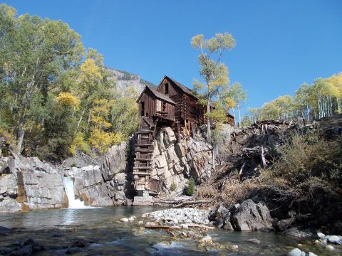 1986 Ford Bronco II - A historic wooden mill structure is perched precariously on a rocky cliff face, with a waterfall cascading into the river below.