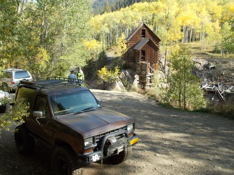 1986 Ford Bronco II - A brown Ford Bronco II is parked on a dirt road with an old wooden structure in the background.