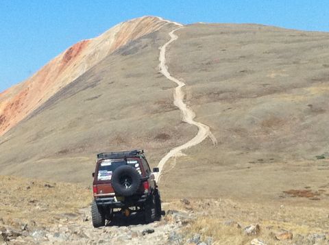1986 Ford Bronco II - A 1986 Ford Bronco II navigates a winding dirt road ascending a barren mountain under a clear blue sky.