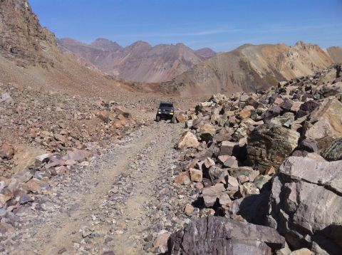 1986 Ford Bronco II - A lone SUV navigates a treacherous, rocky path through a mountainous landscape under a bright blue sky.