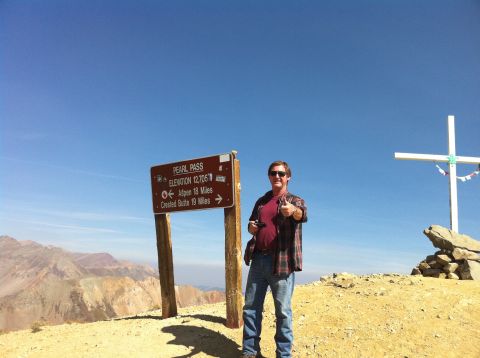 1986 Ford Bronco II - A man stands on a mountain summit near a sign indicating Pearl Pass and its elevation.