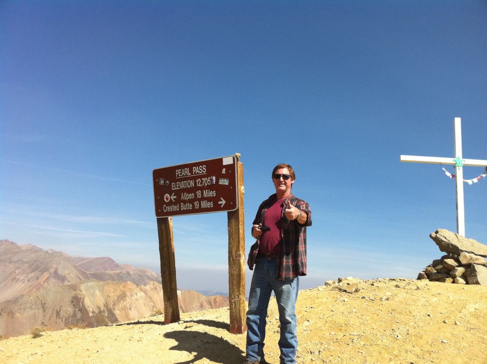 Man in plaid shirt and jeans gives thumbs up near Pearl Pass sign at 12,705 ft elevation