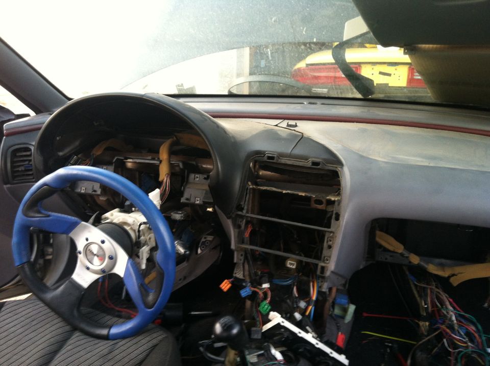 Car interior with disassembled dashboard, blue and black steering wheel, and exposed wiring. Another vehicle is visible through the windshield